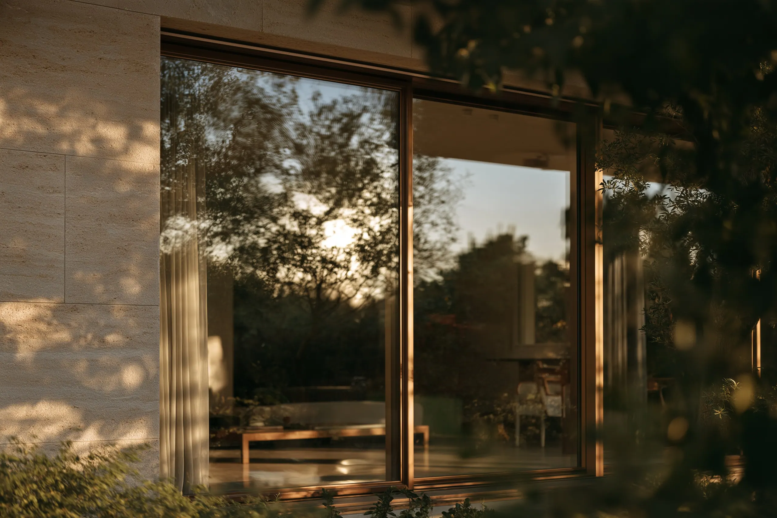 Natursteinfassade mit großer Glasfront, in der sich Bäume im Abendlicht spiegeln